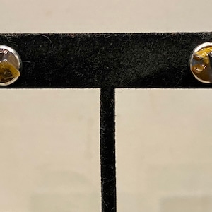 Pode incluir: Um par de brincos circulares com engastes de metal prateado polido. Cada brinco contém fragmentos irregulares incrustados de pedra olho-de-tigre castanho-dourado e peças de pedra cinzento-escura num acabamento de resina transparente e lisa.
