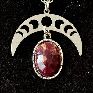 May include: A silver crescent moon necklace with a red gemstone pendant. The moon has cutouts of crescent moons in different phases.