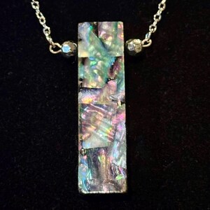Peut inclure: Un collier avec un pendentif rectangulaire et une chaîne argentée. Le pendentif est composé de morceaux irisés en mosaïque dans des tons de vert, de violet et de bleu. Le collier est présenté sur un fond noir.