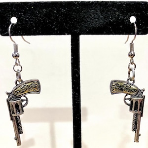 Peut inclure: Une paire de boucles d'oreilles argentées avec des accents dorés, chacune représentant un revolver détaillé.