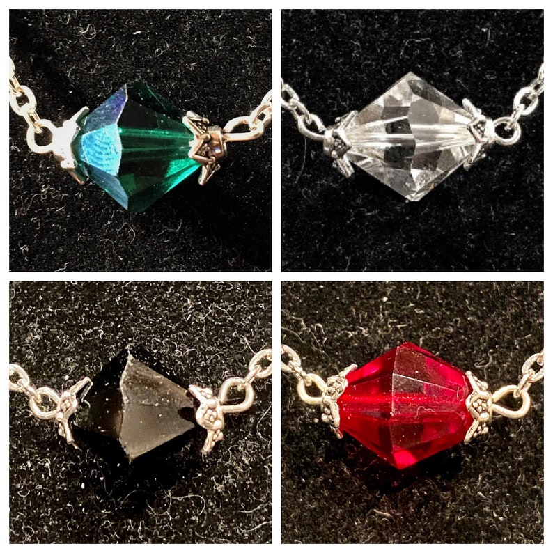 Op de afbeelding: Vier close-up afbeeldingen van zilveren kettingen met verschillende gekleurde kristallen hangers. De hangers hebben de vorm van een diamant. De kleuren zijn groen, helder, zwart en rood.