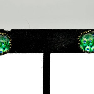 Puede incluir: Un par de pendientes de botón con diseño de pluma de pavo real en un engaste de metal dorado. Los pendientes presentan un diseño de pluma de pavo real verde y azul.