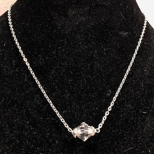 Op de afbeelding: Een zilveren ketting met een heldere kristallen hanger. De hanger heeft een gefacetteerde vierkante vorm.