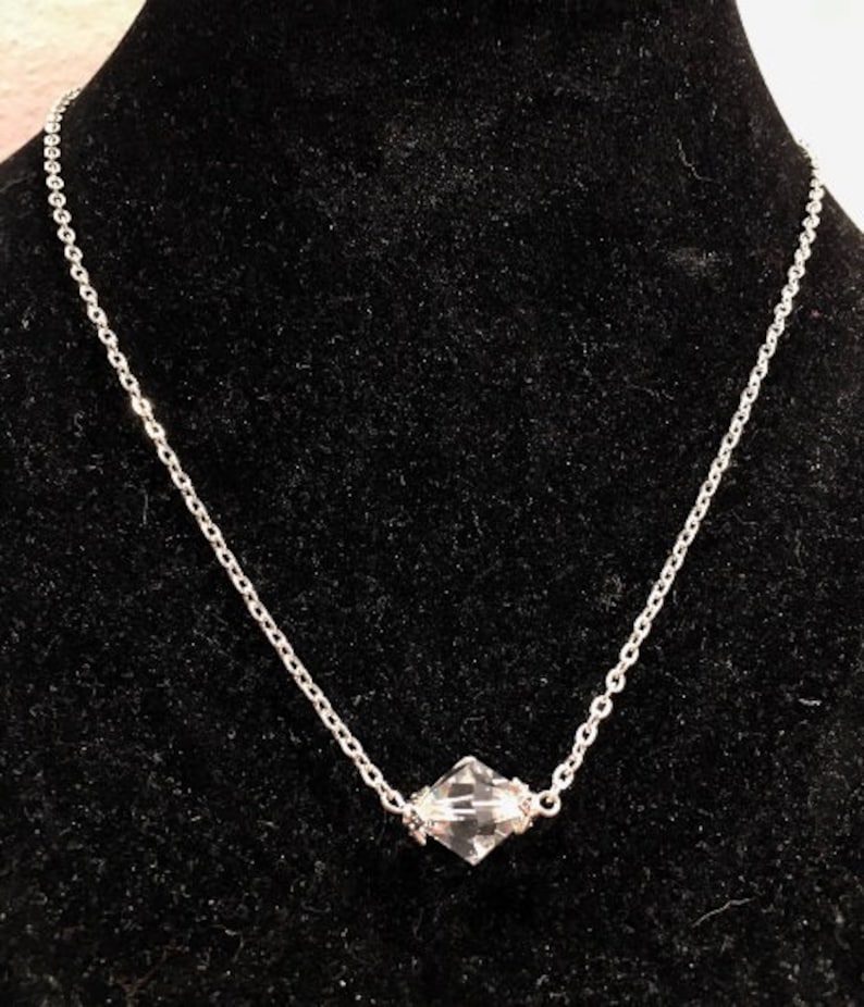 Op de afbeelding: Een zilveren ketting met een heldere kristallen hanger. De hanger heeft een gefacetteerde vierkante vorm.