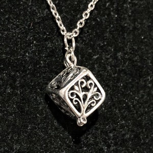 Puede incluir: Un collar de plata con un delicado colgante de cubo adornado y calado. El colgante presenta un diseño floral.