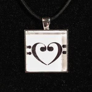 May include: A silver necklace with a black cord and a square pendant featuring a black heart-shaped bass clef on a white background.