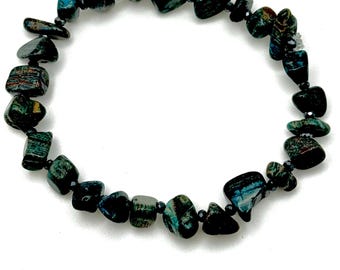 Puede incluir: Un brazalete con piedras pulidas irregulares en tonos turquesa, azul y negro. Las piedras están ensartadas en un cordón oscuro, creando un accesorio flexible. El brazalete está diseñado para adaptarse a una medida de muñeca estándar.