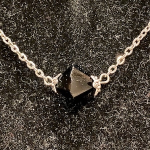Op de afbeelding: Een zilveren ketting met een zwarte edelsteen hanger. De hanger heeft een gefacetteerde, puntige vorm.