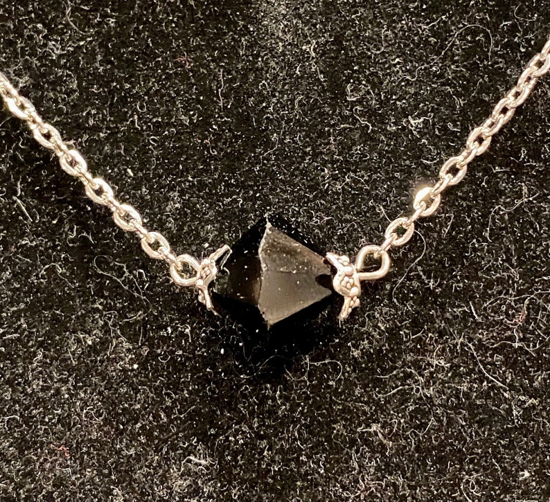 Op de afbeelding: Een zilveren ketting met een zwarte edelsteen hanger. De hanger heeft een gefacetteerde, puntige vorm.