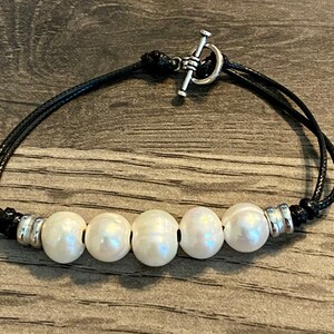 May include: A black cord bracelet with a silver toggle clasp and five white pearls.