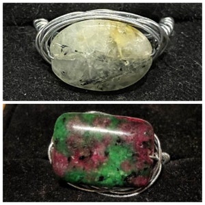 Puede incluir: Dos anillos hechos a mano. El anillo superior presenta una piedra verde claro con inclusiones negras, engarzada en alambre de plata. El anillo inferior muestra una piedra con patrones rojos y verdes, también envuelta en alambre.