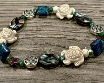 May include: A beaded bracelet featuring three cream-coloured ceramic sea turtle charms, dark blue square beads, iridescent abalone shell oval beads, silver-toned metal filigree discs, small green faceted beads, and dark star-shaped accents.