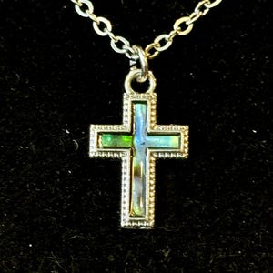 Puede incluir: Un collar de cadena de plata con un pequeño colgante de cruz de plata. La cruz está decorada con ópalo verde y azul iridiscente.