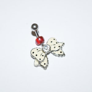May include: A belly button ring featuring a bow design. The bow is off-white with black polka dots and a silver outline. A clear gem is in the center of the bow. The ring has a silver barbell and a red bead.