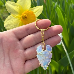 May include: A handmade keychain with a light blue, iridescent arrowhead-shaped crystal wrapped in gold-coloured wire. The keychain has a gold-coloured ring and chain. The background includes a yellow daffodil and green foliage.