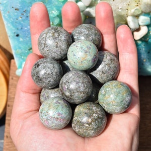 Peut inclure: Une collection de sphères de pierres précieuses vertes et grises, également connues sous le nom d'aventurine verte, dans une main. Les sphères sont lisses et polies.