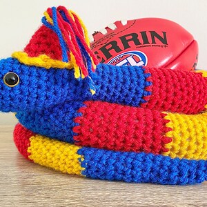 Op de afbeelding: Een gehaakt slangen speelgoed in blauw, rood en geel, met een rode en witte Australische voetbal op de bovenkant. De voetbal heeft het woord "ERRIN" erop gedrukt.