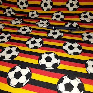 Könnte beinhalten: Schwarz, rot und gelber gestreifter Stoff mit einem sich wiederholenden Muster aus weißen Fußbällen.