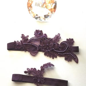 May include: A set of two dark purple elastic garters featuring intricate lace floral appliqués embellished with matching dark purple beads. Above the garters sits a faceted, clear crystal-like circular ornament with the word BRIDE etched in gold.