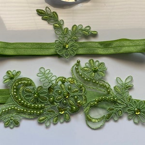 May include: Two green elastic garters with delicate green lace and pearl embellishments. The lace features floral and leaf designs.