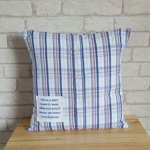 May include: A blue and red plaid pillow made from a repurposed shirt. The pillow has a small white fabric patch sewn on the side with the text "This is a shirt I used to wear, when you hold it know I am there. Love Dad xxx".