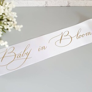 May include: White satin sash with gold lettering that reads "Baby in Bloom".