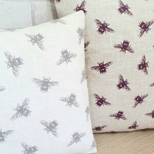 May include: Two decorative throw pillows with a beige linen fabric and a repeating pattern of bees. One pillow has a gray bee pattern, and the other has a burgundy bee pattern.