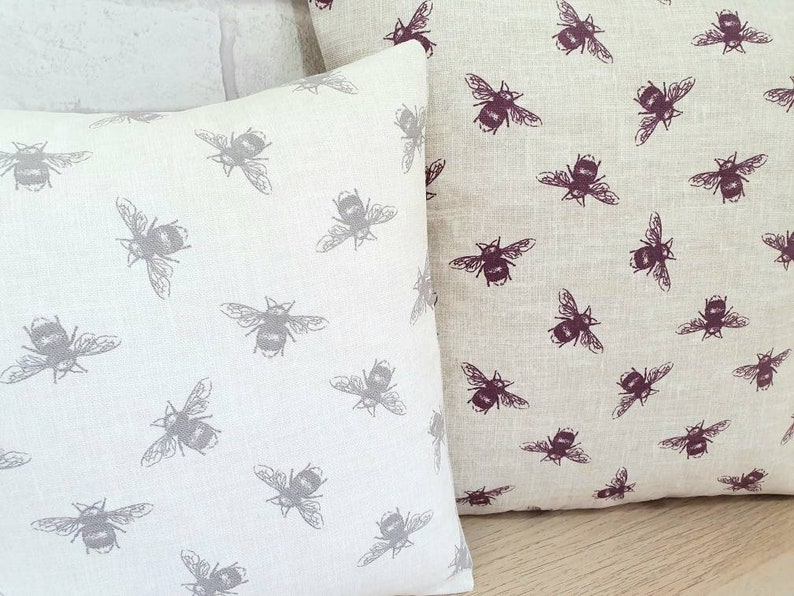 May include: Two decorative throw pillows with a beige linen fabric and a repeating pattern of bees. One pillow has a gray bee pattern, and the other has a burgundy bee pattern.