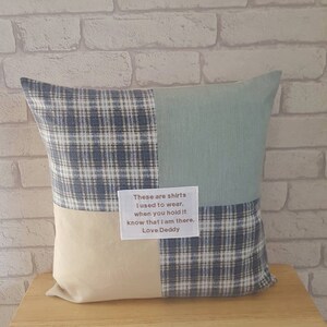 May include: A square pillow with a patchwork design. The pillow is made of four different fabrics: a blue and white plaid, a light blue solid, a beige solid, and another blue and white plaid. The pillow has a white label with the text "These are shirts I used to wear, when you hold it know that I am there. Love Deddy."