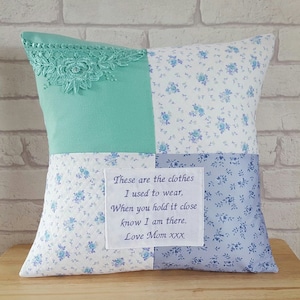 May include: A square patchwork cushion with a light blue floral pattern on three sides. The fourth side is a light green fabric with a white lace trim. A white fabric label with a message is sewn onto the cushion: "These are the clothes I used to wear, When you hold it close know I am there. Love Mom xxx".