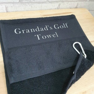 May include: Black golf towel with a silver carabiner clip. The towel has the text "Grandad's Golf Towel" embroidered on it in white.
