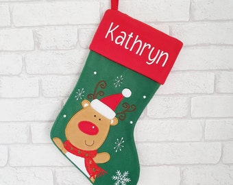 May include: A Christmas stocking with a red cuff and a green body. The word "Kathryn" is embroidered in white on the cuff. A reindeer wearing a Santa hat and scarf is on the front, with snowflakes.