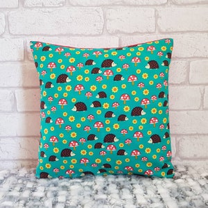 May include: A turquoise blue throw pillow with a repeating pattern of brown hedgehogs, red mushrooms, and yellow flowers.