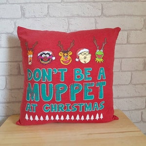 May include: Red Christmas pillow with a green and white "Don't be a Muppet at Christmas" message. The pillow features a variety of Muppet characters, including Animal, Miss Piggy, Fozzie Bear, Gonzo, and Kermit the Frog.