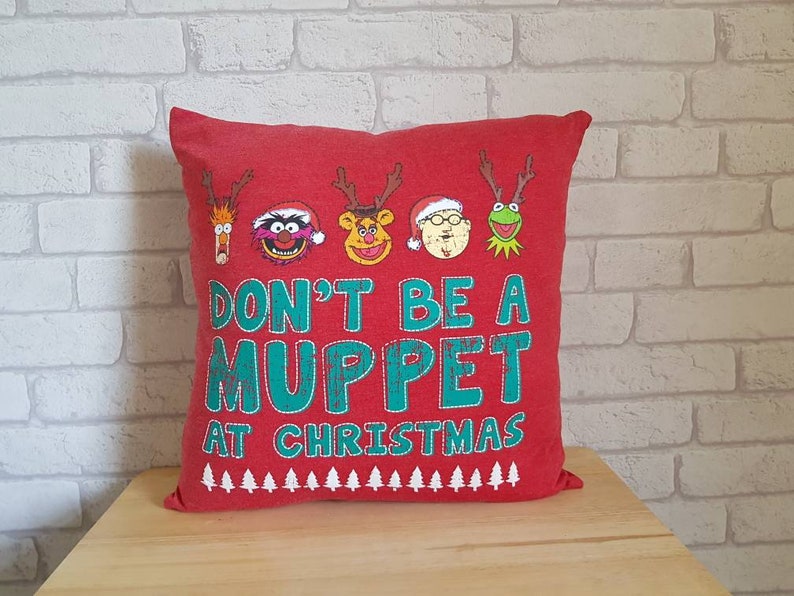 May include: Red Christmas pillow with a green and white "Don't be a Muppet at Christmas" message. The pillow features a variety of Muppet characters, including Animal, Miss Piggy, Fozzie Bear, Gonzo, and Kermit the Frog.