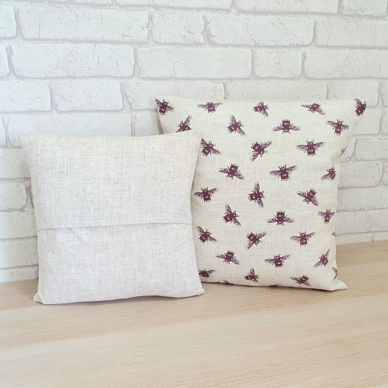 May include: Two throw pillows, one with a beige linen fabric and the other with a beige linen fabric with a repeating pattern of dark purple bees.