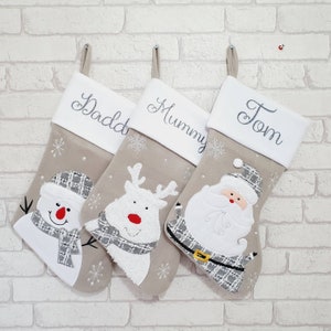 May include: Three personalized Christmas stockings with gray and white fabric. The stockings feature a snowman, reindeer, and Santa Claus. The stockings are embroidered with the names "Daddy", "Mummy", and "Tom".