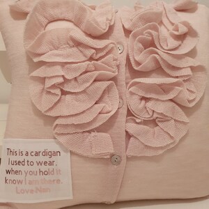 May include: A pink knitted cardigan pillow with a ruffled collar and buttons. The pillow has a white fabric label with the text "This is a cardigan I used to wear. When you hold it know I am there. Love Nan."