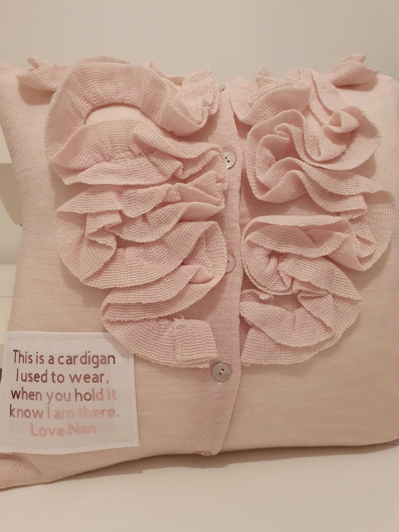 May include: A pink knitted cardigan pillow with a ruffled collar and buttons. The pillow has a white fabric label with the text "This is a cardigan I used to wear. When you hold it know I am there. Love Nan."