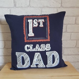 May include: A navy blue throw pillow with white text that reads "1st Class Dad" in a chalkboard style font. The text is framed in a red and white border.