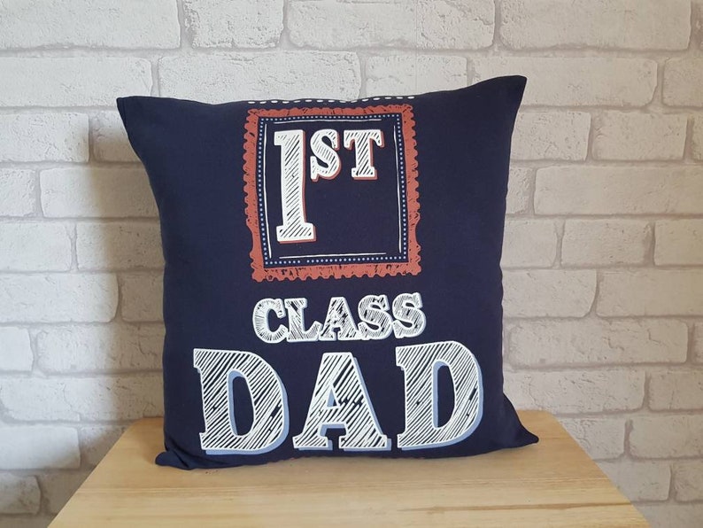 May include: A navy blue throw pillow with white text that reads "1st Class Dad" in a chalkboard style font. The text is framed in a red and white border.