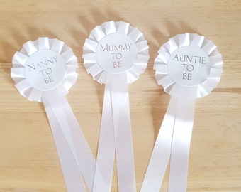 May include: Three white satin rosettes with white text on a white background. The rosettes read "Nanny to Be", "Mummy to Be", and "Auntie to Be".