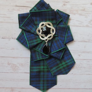 Könnte beinhalten: Eine Schottenmuster-Brosche in Blau, Grün und Schwarz mit einem silberfarbenen Mittelstück und einem schwarzen Tropfenanhänger. Die Brosche ist aus Stoff gefertigt und hat ein geschichtetes Design.