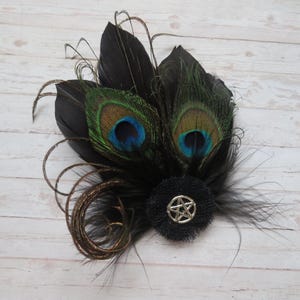 May include: A decorative hair clip featuring black feathers and two peacock feathers with iridescent blue and green eye markings. A black fabric flower with a silver pentagram is at the center, with gold and black accents.