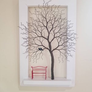 May include: A white framed wall art piece featuring a wire sculpture of a bare tree with two black birds perched on a branch. A red wire bench sits below the tree on a small shelf.