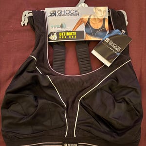 May include: Black sports bra with white trim and the text "SHOCK ABSORBER". The bra has a mesh panel and thick straps. The packaging includes a picture of a woman wearing the bra and the words "ULTIMATE RUN BRA".