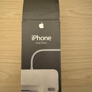 Può includere: Scatola nera per iPhone Dual Dock con il logo Apple. La scatola presenta le parole "iPhone Dual Dock" in bianco. Il dock stesso è bianco con un cavo bianco.