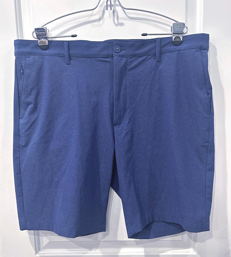 May include: Blue shorts with a textured pattern, featuring a button closure and pockets. The shorts are hanging on a silver hanger. The shorts are a solid blue color.