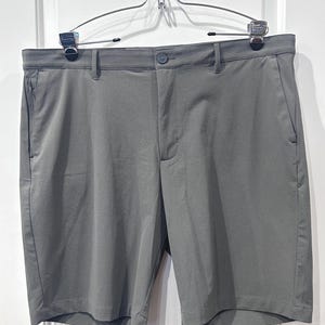 May include: Gray shorts with a button closure, belt loops, and side pockets. The shorts are hanging on a metal hanger against a white background. The fabric has a subtle textured pattern.
