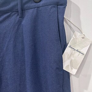 May include: Close-up of navy blue pants with a textured pattern. The pants have a pocket and a tag that reads "Tommy Bahama". The pants are hanging on a hanger.